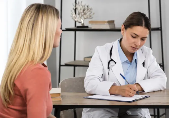 Médica de jaleco branco anota informações em uma prancheta enquanto atende uma paciente sentada à sua frente, em consultório clínico.