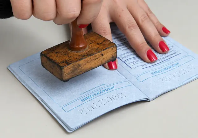 Mãos carimbando uma carteira de trabalho aberta sobre a mesa, com unhas vermelhas e páginas azuis visíveis.